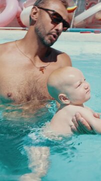 Young father carefully holding his adorable baby in a swimming pool for the first time, enjoying a fun family activity and bonding together on a sunny summer day during their vacation. Vertical video