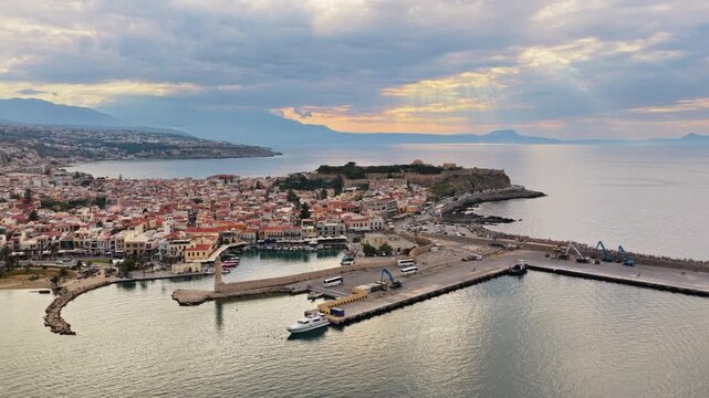 Aerial drone view of a long sandy town beach with straw parasols along the waterfront, backed by hills, Rethymno, Greece
