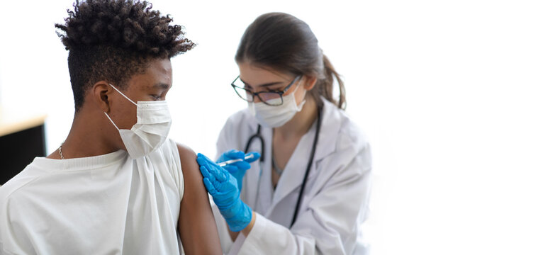 asian woman doctor holding syringe and injection vaccine shot to black male patient. Young african american man sitting and getting vaccinated against coronavirus in hospital