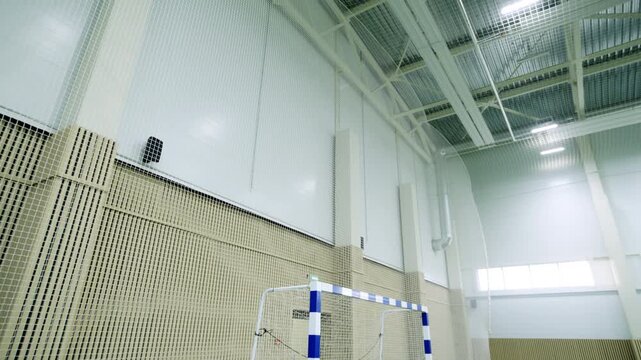 Goal with balls stands on green turf pitch in empty sports hall. Bright overhead lights illuminate vast silent space before game starts
