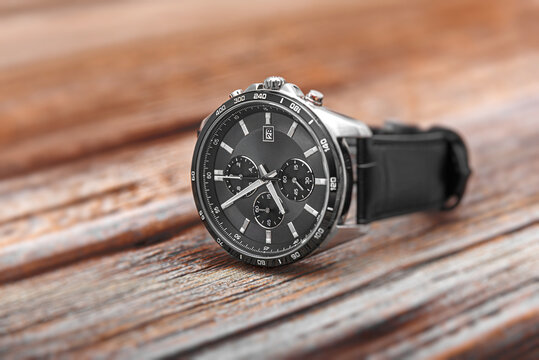 A modern silver chronograph wristwatch with a black dial and black leather strap lies on a rustic wooden surface.