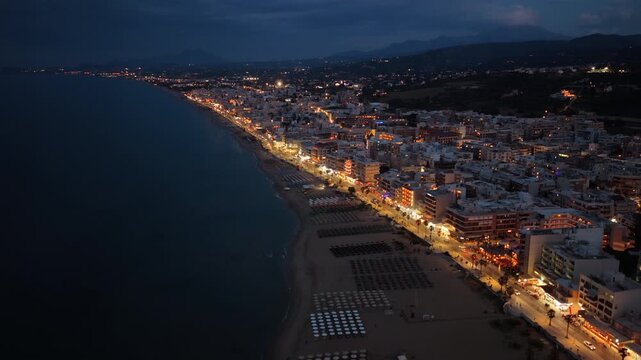 Aerial drone view of the long sandy town beach at night below the illuminated seafront promenade, Rethymno, Greece