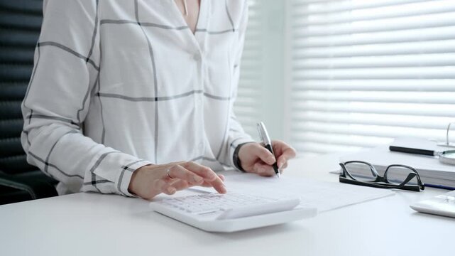 Businesswoman hands calculating finance, bookkeeping, and taxes, using an office calculator while writing down important financial data on documents at a desk