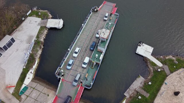 Aerial drone view of vehicle ferry transporting cars across a wide river with automobiles boarding and disembarking at ferry terminal connecting opposite riverbanks transportation infrastructure RO-RO