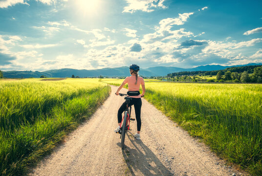 Woman riding mountain bike on dirt road through green wheat fields at sunset in Slovenia. Female cyclist cycling in countryside landscape and sunny sky. Summer outdoor activity, fitness. Girl on bike