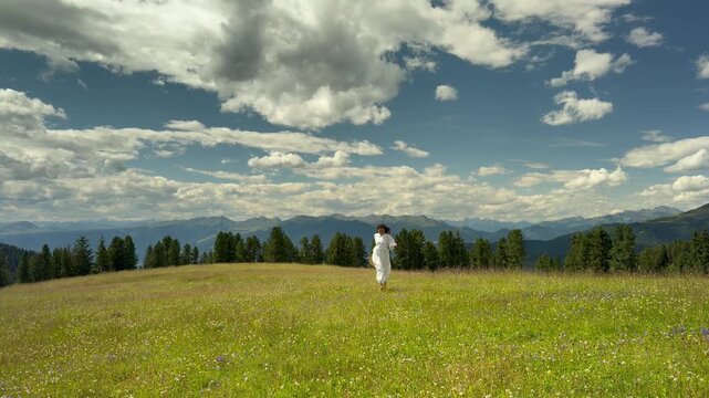 Beautiful Carefree Woman in a Flowing White Dress and Straw Hat Running Freely in a Wide Alpine Meadow With the Iconic Odle Geisler Spitzen Dolomite Peaks and Dramatic Cloudy Sky, Val Gardena South Ty