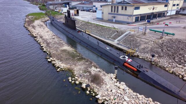 Aerial view of a submarine museum display beside a river, with the black submarine moored in a narrow channel near industrial buildings and rock-lined banks