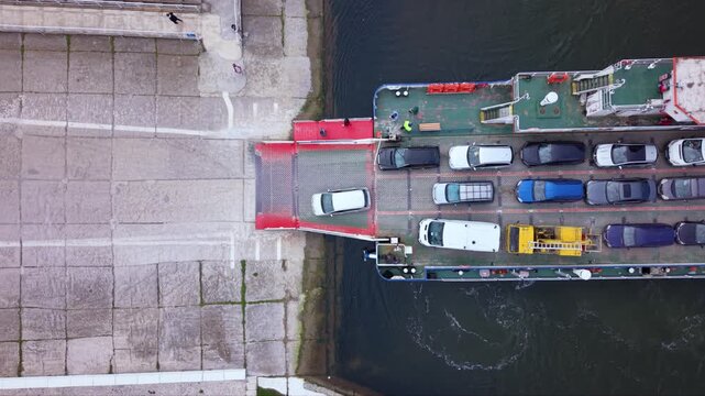 Aerial drone view of vehicle ferry transporting cars across a wide river with automobiles boarding and disembarking at ferry terminal connecting opposite riverbanks transportation infrastructure RO-RO