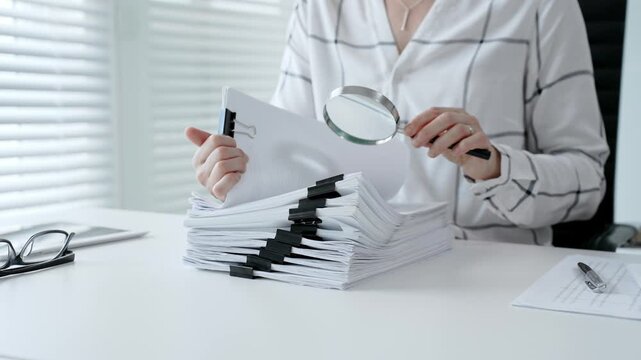 Professional woman investigating important business documents, scanning legal papers with a magnifier, performing a thorough audit, analyzing reports and financial data