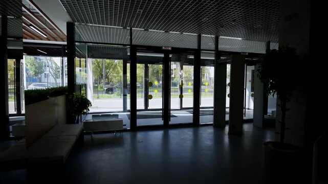 Empty lobby features glass entrance doors with caution circles. Dark reflective floor leads past potted plant towards tables and chairs