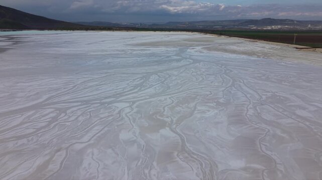 Aerial drone footage flying low above white mineral evaporation pond with intricate natural patterns and textured sediment surface in industrial extraction area surrounded by hills dramatic cloudy sky