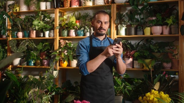 Portrait of modern professional caucasian man working as florist using smartphone online at flowers shop communicating with customers. Beautiful young adult male worker taking care of flowers.