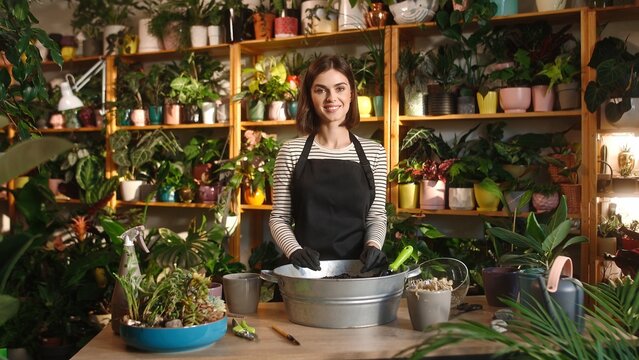 Portrait young successful attractive woman mixing soil for with small shovel working at beautiful flower shop. Pretty cute adult female florist wearing apron looking at camera smiling.