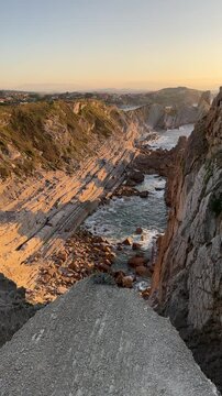 Walking along a narrow rocky ledge at the cliff edge overlooking deep coastal rock fissures and the Costa Quebrada coastline in warm golden evening light.