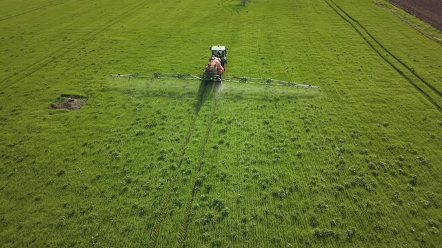 Aerial view of agricultural spraying on a lush green field, with a tractor pulling a wide boom sprayer across crop rows