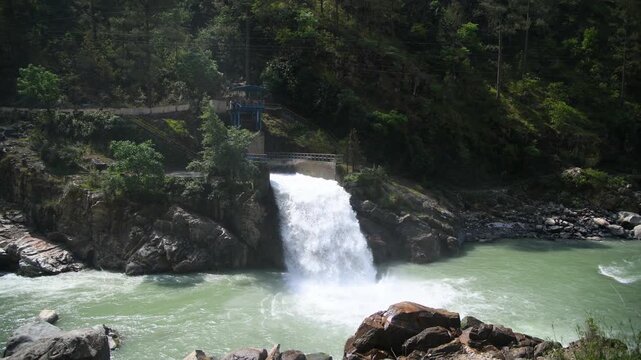 A scenic wide-angle shot showing a powerful gush of white water discharging into the turquoise green Bhagirathi River near Maneri Dam (Maneri Bhali Pariyojna Stage-1) in Uttarkashi, Uttarakhand