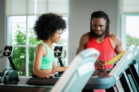 Young girl focused intently while an adult man guides her on a treadmill. He points at the display, smiling encouragingly. She listens carefully, showing determination and learning