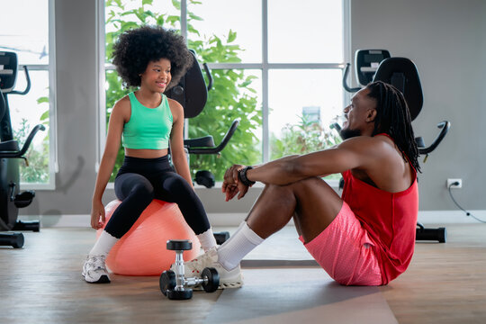 Black fitness coach dad smiling teen girl resting fitness ball. Scene for family wellness, personal trainer mentoring child, cardio weight training break and communication