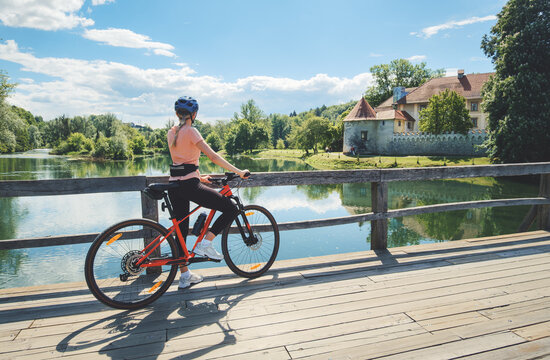 Female cyclist with mountain bike standing on wooden bridge in Slovenia. Woman looking at historic Otocec Castle on Krka river under blue sky. Summer cycling trip, travel destination. Girl on bike