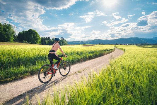Woman riding mountain bike on dirt road through green wheat fields at sunset in Slovenia. Female cyclist cycling in countryside landscape and sunny sky. Summer outdoor activity, fitness. Girl on bike
