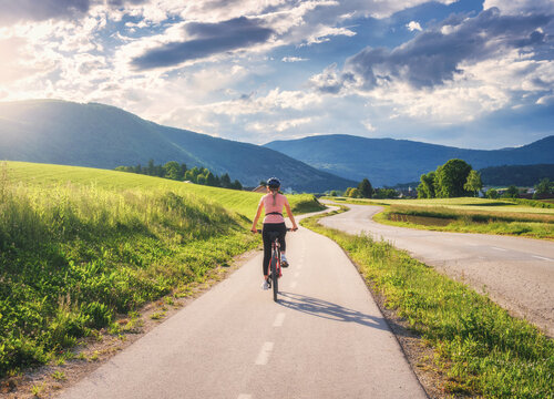 Woman riding mountain bike on asphalt bicycle path in Slovenia countryside in summer. Female cyclist cycling on lane through green meadows and hills at sunset. Summer outdoor activity, fitness. Travel