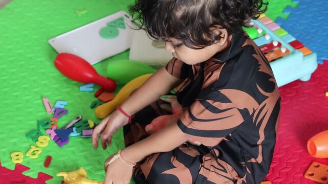 High angle view of a cute Indian toddler girl sitting on a colorful playmat indoors, deeply engaged in unstructured independent play with various scattered educational toys, letters, and animal figure