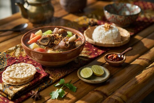 Traditional indonesian sup buntut oxtail soup served in rustic clay bowl with steamed white rice side dish and lime wedges on wooden bamboo table setting.