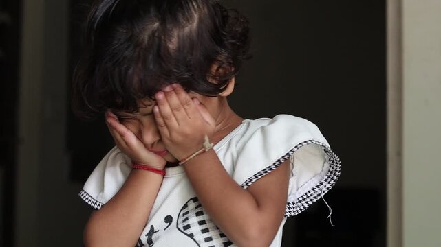 Close-up portrait of a cute little Indian toddler girl covering her face with her hands, looking shy, sad, or upset against a dark background.
