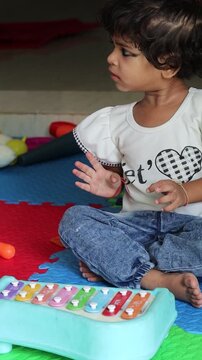 Vertical candid shot of a cute little Indian toddler girl sitting on a colorful play mat at home, playing with a vibrant musical xylophone toy.
