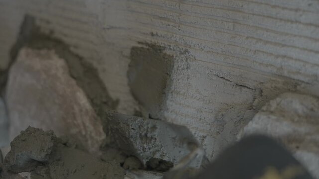 Point-of-view shot of a mason's hands using a spatula trowel to apply wet mortar paste onto a stone wall structure. 