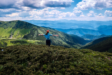 Obraz premium female hiker standing on a lush green mountain summit with her arms spread wide, embracing the freedom and the breathtaking panorama of layered mountain ridges and valleys under a cloudy sky