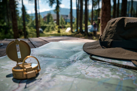A horizontal low-angle shot of an open golden military compass and a khaki safari hat resting on a paper map spread across an outdoor table, with a blurred background of a pine forest camping ground