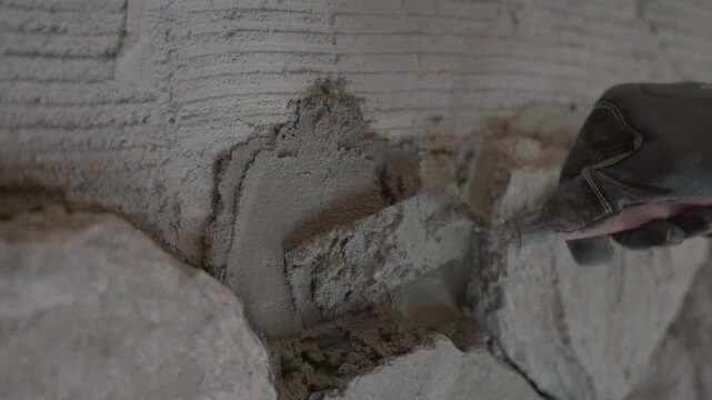 Close-up shot of a hand in a protective construction glove using a trowel to apply and smooth wet mortar paste between stones on a wall.