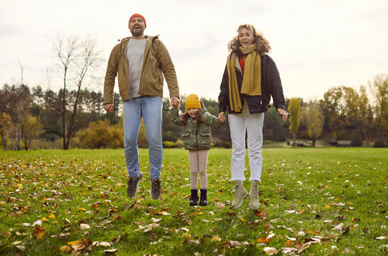 Family parents child autumn park jumping together. Cheerful mother and father hold hands with their girl, enjoying colorful leaves and fresh air. Happy family autumn joy and togetherness.