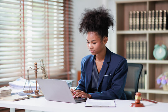 Young female lawyer or legal professional working on laptop computer in modern office, managing legal documents and representing justice with scales and gavel on desk