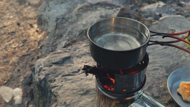 Water boiling in steel pot on wood-burning camping stove with glowing embers, outdoor survival cooking scene