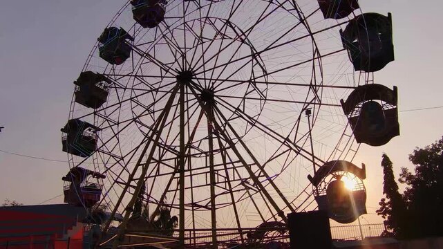 Silhouetted Ferris wheel spins against twilight sky. Sun sets behind amusement park ride, casting warm glow. Passengers ride, suspended in slow, rhythmic motion. Structure stands tall