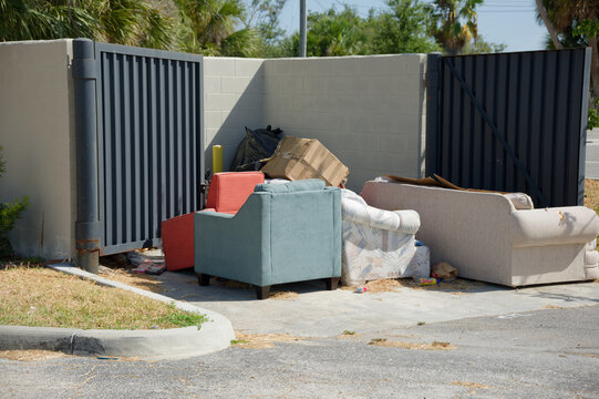 Discarded sofas and armchairs sit beside a metal dumpster enclosure at a residential curb, surrounded by cardboard and trash, suggesting illegal dumping, neglect and bulky furniture disposal on a sunn