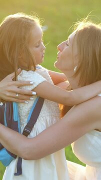 Young daughter with a backpack hugging and kissing her smiling mother goodbye before school, sharing a tender and loving moment together outdoors in the warm sunset light against a green background