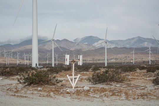 A small white weather monitoring station stands in a desert field with massive wind turbines and rocky mountains behind.