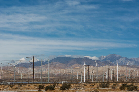 Rows of spinning wind turbines at a desert facility framed by multiple horizontal electrical power lines and a giant mountain range.