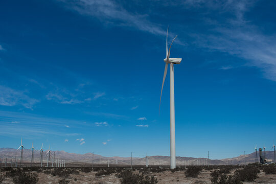 Low angle view of a single massive modern wind turbine propeller spinning against a bright blue sky with wispy white clouds.