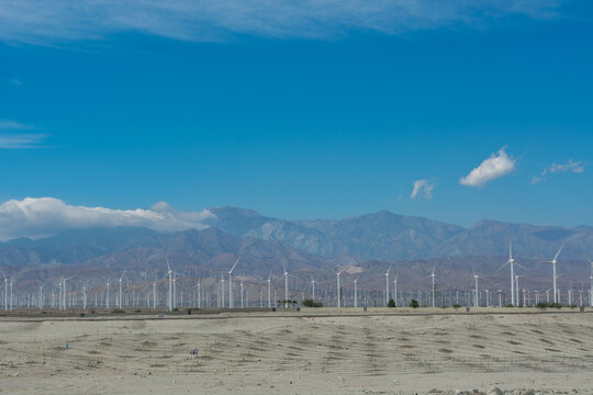 Hundreds of wind turbines stretching across a dry desert valley with power lines and majestic mountains in the distance.