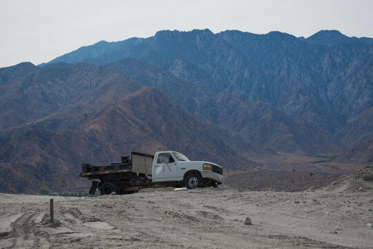 Old white dump truck parked on a sandy desert hill with a massive rugged mountain range in the background.