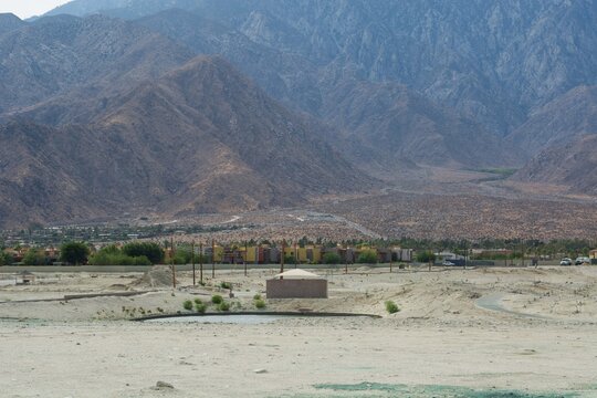 Wide landscape view of a small desert oasis pond with a small building at the foot of massive rocky mountains.