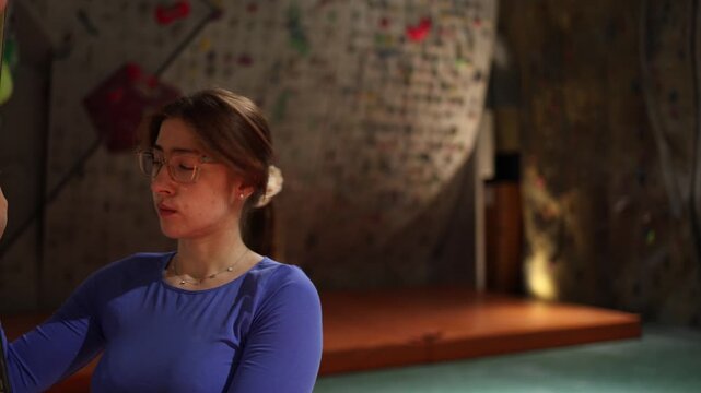 Beginner woman on indoor climbing wall carefully checks safety rope before next move showing focus preparation and attention during first training session