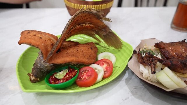 Fried gourami and crispy grilled chicken served with fresh chili sauce, cucumber, and tomato on a green plate, a traditional Indonesian dish perfect for culinary and restaurant video shoots.