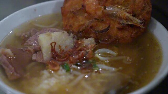 Traditional Indonesian beef soup with rice, bean sprouts, fried shallots, herbs, and crispy crackers, captured in close-up culinary footage.