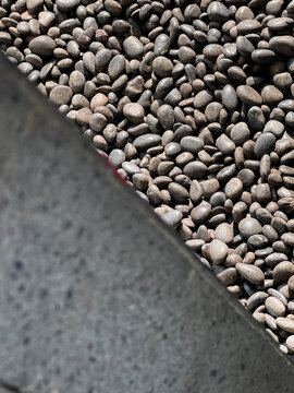 Diagonal composition of smooth dark river pebbles bordering a textured gey concrete curb, creating a natural geometric contrast between organic stone and man-made material.