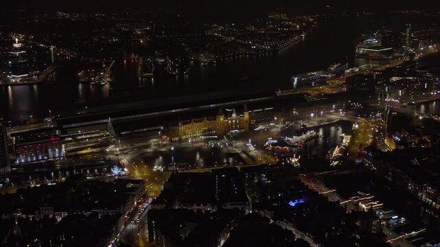 Aerial night view of Amsterdam Central Station, Netherlands, with the illuminated railway terminal, ferry docks on the IJ river, canal bridges, holiday lights and traffic in the city center.